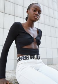 Top cropped preto com nervuras, detalhe de nó frontal e lados abertos, combinado com jeans brancos de cintura alta e um cinto preto com tachas.