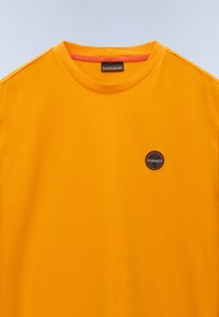 Camiseta de algodón amarilla con cuello redondo y un pequeño logo negro redondo en el lado izquierdo del pecho. Textura suave y mangas cortas.