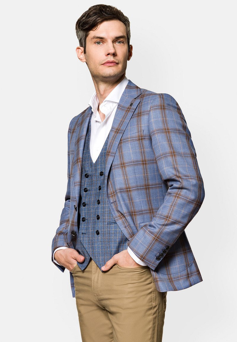 Niebieska marynarka w kratę z brązowymi akcentami, zestawiona z niebieską kamizelką w zygzaki, białą koszulą i khaki spodniami. Dopasowany krój i teksturowana tkanina.
