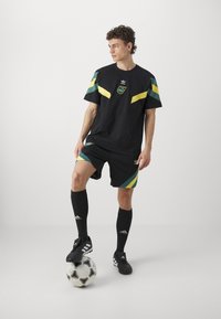 Camiseta de fútbol negra con acentos verdes y amarillos, combinada con pantalones cortos negros con rayas diagonales. Calcetas negras y botas de fútbol.