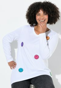 Glimlachende vrouw met krullend haar zit, gekleed in een witte lange mouwen shirt versierd met kleurrijke smiley patches en donkere spijkerbroek.