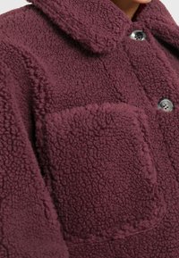 Primer plano de una chaqueta de forro polar burdeos con un gran bolsillo de parche, tejido texturizado y botones grises brillantes cerca del cuello.