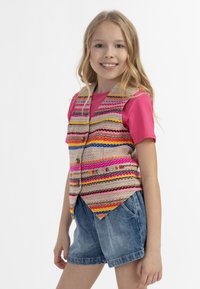 Spalvingas audinio liemenė su daugybe spalvotų horizontalių juostelių, mygtukai priekyje, nešiojama virš rožinės marškinės ir derinama su džinsiniais šortais.