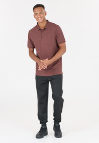 Burgundyröd kortärmad polo med ribbad krage, tillsammans med svarta tapered byxor och svarta sneakers. Modellen står med händerna ihop.