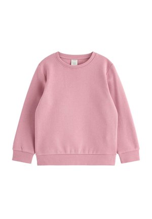 Ung pige med krøllet hår har en lyserød sweatshirt på med mønstrede lange ærmer, mørke bukser og sko, og står med benene bredt fra hinanden på en hvid baggrund.