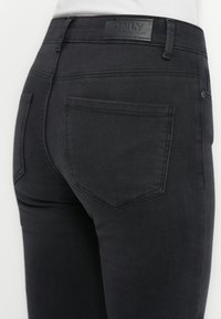Mustad denim püksid, millel on keha järgiv lõige, sile tekstuur, kaks tagumist taskut ja logo silt vöökoha peal.