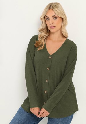 Blonde Frau trägt ein olivgrünes, geknöpftes Langarmshirt und blaue Jeans, posiert vor einem schlichten weißen Hintergrund.