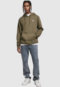 Bluza z kapturem w oliwkowej zieleni, wykonana z miękkiego materiału, z kangurową kieszenią i regulowanym sznurkiem. Nosi się ją z jasnoniebieskimi jeansami o prostych nogawkach i białymi trampkami.