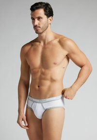 Weiße Herren-Bikiniunterwäsche mit silbernen Akzenten, elastischer Taillenbund mit dem Schriftzug "HUMAN", enganliegendes Design und glatte Textur.