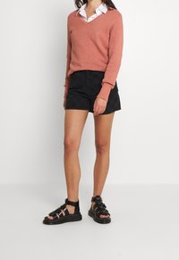 Hellkorallenfarbener Strickpullover mit V-Ausschnitt, kombiniert mit schwarzen karrierten Shorts. Schwarze Riemchensandalen mit Nieten vervollständigen das Outfit.