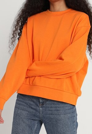Personne aux longs cheveux bouclés porte un pull oversize orange vif et un jean bleu sur un fond uni.