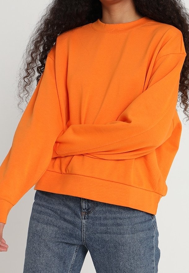 Personne aux longs cheveux bouclés porte un pull oversize orange vif et un jean bleu sur un fond uni.