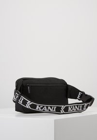 Schwarze Stoff-Schultertasche mit Reißverschluss, verstellbarem Trageriemen mit weißen Buchstaben, rechteckiger Form, minimalistischem Design und glatter Textur.