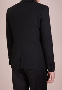 Blazer nero su misura con una texture liscia, apertura posteriore singola e spalle strutturate. Presenta bottoni neri sui polsini delle maniche.