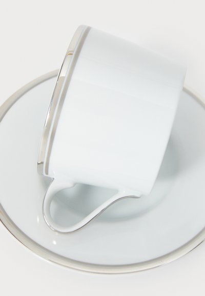 Biały ceramiczny kubek i spodek z srebrną obręczą, o gładkiej fakturze i zakrzywionym uchwycie, ustawione pod kątem.