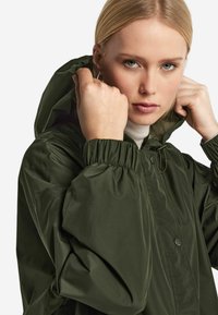 Olivgrön zip-up jacka med huva, elastiska ärmslut och en slät, glänsande yta. Har knappar och sitter tajt runt ansiktet.