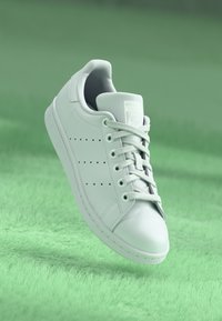 Bílé tenisky Adidas Stan Smith s tkaničkami na zeleném texturovaném povrchu, nakloněné tak, aby byly jasně vidět detaily z boku a shora.
