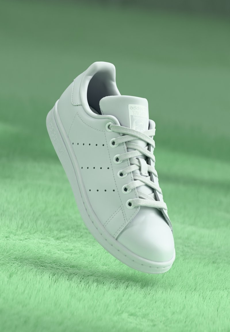 Bílé tenisky Adidas Stan Smith s tkaničkami na zeleném texturovaném povrchu, nakloněné tak, aby byly jasně vidět detaily z boku a shora.