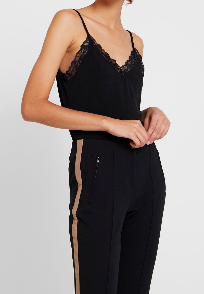 Camisole noir avec des bordures en dentelle et des bretelles fines, associé à un pantalon noir taille haute avec une bande latérale beige et des poches zippées.