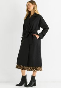 Black trench coat with a belted waist, featuring a collar and side pockets, layered over a leopard print dress. Worn with black ankle boots.