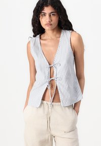 Top senza maniche bianco e blu a righe, con scollo a V profondo, fronte annodato e vestibilità ampia. Abbinato a pantaloni beige chiaro con cordino.