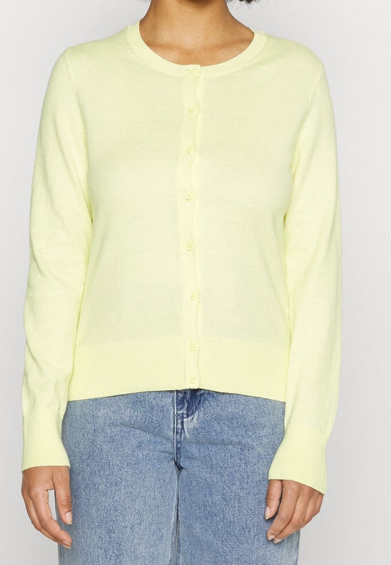 Femme portant un cardigan boutonné jaune pâle et un jean taille haute bleu clair, visible du cou aux hanches sur un fond uni.