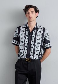 Camisa de manga corta negra con un patrón de cadena plateada, diseñada con un cierre de botones en la parte delantera y un corte relajado, combinada con pantalones negros.