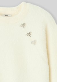 Pull couleur crème avec une texture douce et duveteuse, orné de trois embellissements en forme de nœuds tons argentés. Encolure ronde et poignets côtelés.