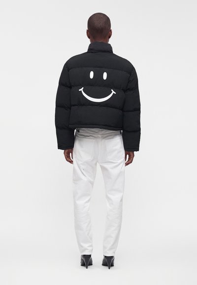 Personne portant une doudoune noire avec un grand visage souriant blanc dans le dos, un pantalon blanc et des chaussures à talons noires, vue de dos.