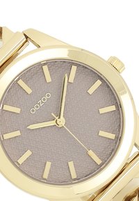 Montre bracelet en or avec un cadran gris texturé, des index et aiguilles en or. Elle dispose d'un bracelet en métal poli et d'une couronne sur le côté.