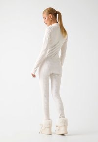 Combinaison intégrale blanche au design lisse et ajusté, avec une texture subtilement structurée. Des bottes douces, duveteuses et de couleur crème complètent la tenue.