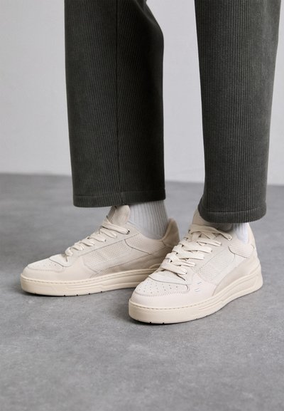 Beżowe sneakersy wykonane z zamszu i siateczki, z perforowanymi akcentami, płaskimi sznurówkami i grubą podeszwą. Noszone z zielonymi spodniami z korduropy.