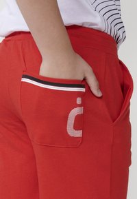Pantaloni felpati rossi con tasca laterale, caratterizzati da un bordo a righe bianche e nere e un elemento grafico a contrasto. Materiale in cotone morbido.