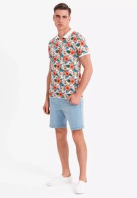 Blommig polo-shirt med orange och vita blommor, gröna blad och en vit krage, matchad med ljusblå denimshorts och vita skor.