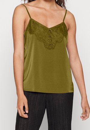 Mujer con camisola de satén verde oliva con detalles de encaje y tirantes finos, combinada con pantalones plisados negros sobre un fondo liso.