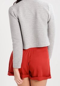 Veste courte grise côtelée associée à un short rouge, avec une poche arrière et un ourlet retroussé. Textures lisses avec des couleurs contrastées.