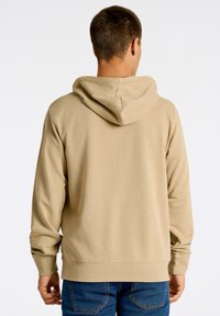 Felpa beige con cappuccio regolabile, polsini a coste e tessuto testurizzato. Indossata con jeans blu, vista da dietro.