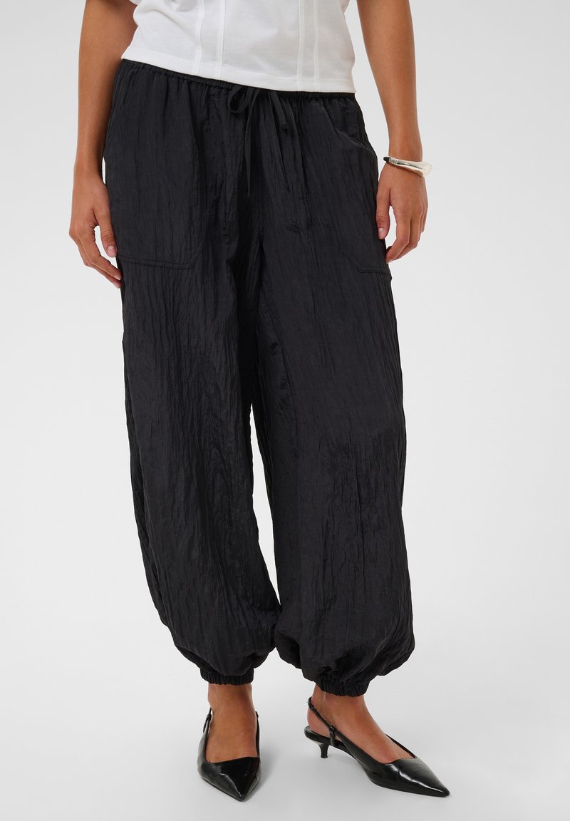 Femme portant un pantalon noir ample froissé avec des poignets élastiques, un haut blanc, des escarpins noirs pointus à bride arrière et un bracelet argenté au poignet droit.