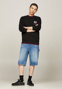 Sudadera negra con detalles rojos, pantalones cortos de mezclilla azules, zapatillas negras con detalles blancos y calcetines negros. Logo en el lado izquierdo del pecho.