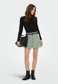 Femme en haut noir à manches longues, jupe en velours côtelé verte boutonnée avec ceinture, bottines noires, tenant un sac à main noir sur son bras gauche.