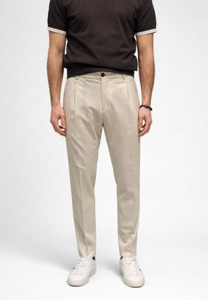 Uomo con pantaloni beige plissettati, sneakers bianche, bracciale nero e camicia nera a maniche corte su sfondo semplice.