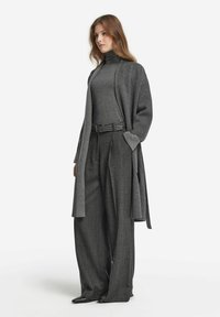 Grau gestrickte Strickjacke über einem grauen Rollkragenpullover, kombiniert mit weit geschnittenen, strukturierten schwarzen Hosen. Enthält einen taillierten Gürtel und spitze Schuhe.