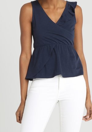 Haut cache-cœur sans manches bleu marine avec détail volanté à l'épaule, associé à un pantalon blanc ajusté porté par une personne debout.