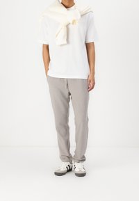 Persoon in een wit t-shirt, lichtbeige sweatpants, witte sneakers met zwarte strepen en een lichtbeige sweatshirt over de schouders.