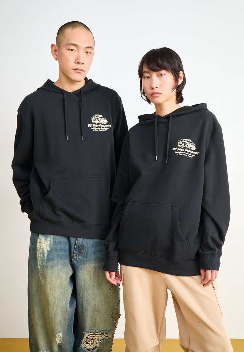 Black hooded sweatshirts with front pockets, featuring a logo on the chest. Worn with distressed jeans and beige loose-fitting pants.