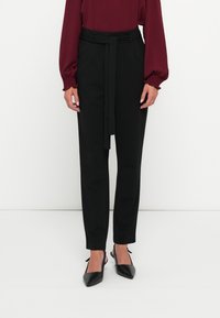 Schwarze maßgeschneiderte Hose mit tailliertem Bund, schmal zulaufendem Bein und glatter Textur, kombiniert mit spitzen schwarzen Absätzen und einer burgunderfarbenen Bluse.