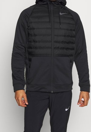 Homme portant une veste à capuche zippée noire Nike avec un panneau avant matelassé et un pantalon noir Nike assorti avec logo blanc.