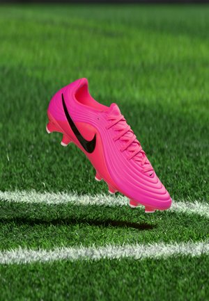 Chaussure de football Nike rose vif avec logo swoosh noir flottant au-dessus d'une pelouse verte marquée de lignes blanches.