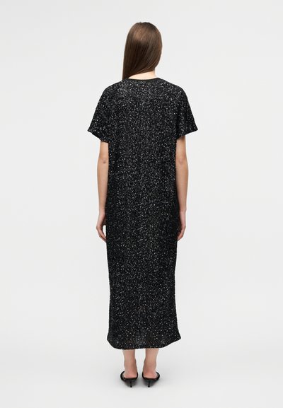 Μαύρο μάξι φόρεμα με κοντά μανίκια, κατασκευασμένο από υφασμάτινο ύφασμα με κηλίδες και υφή. Ο σχεδιασμ ός είναι φαρδύς και φτάνει μέχρι τους αστραγάλους, συνοδευόμενος από παπούτσια με χοντρό τακούνι.