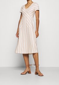 Abito a righe in beige chiaro con sottili linee marroni, maniche corte, scollatura a V e due tasche frontali, abbinato a sandali marroni con cinturini.